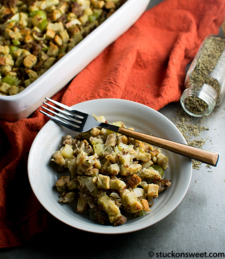 Golden baked stuffing with celery and herbs served on white plate, side view, with kitchen herbs and baking dish in background, perfect for holiday dinners and stuffing recipes.