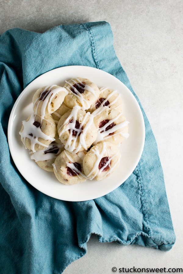 Cream cheese-filled cookie sandwiches drizzled with white chocolate and topped with fruit preserves on a white plate over a blue napkin.