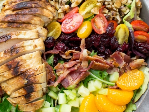 Grilled chicken salad with mixed greens, cherry tomatoes, mandarin oranges, bacon, dried cranberries, chopped cucumbers, walnuts, and ranch dressing on a white plate.