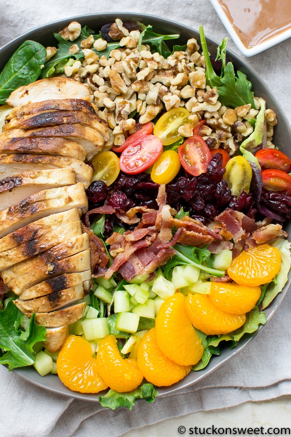 Grilled chicken salad with mixed greens, cherry tomatoes, mandarin oranges, bacon, dried cranberries, chopped cucumbers, walnuts, and ranch dressing on a white plate.