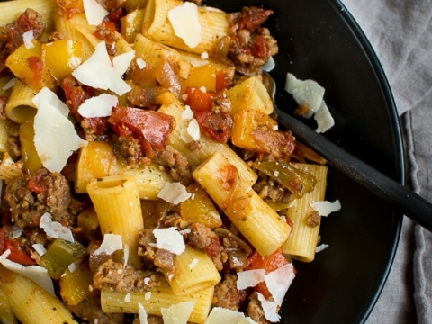 Sweet Italian sausage baked ziti with colorful peppers and shaved Parmesan cheese in a black bowl.