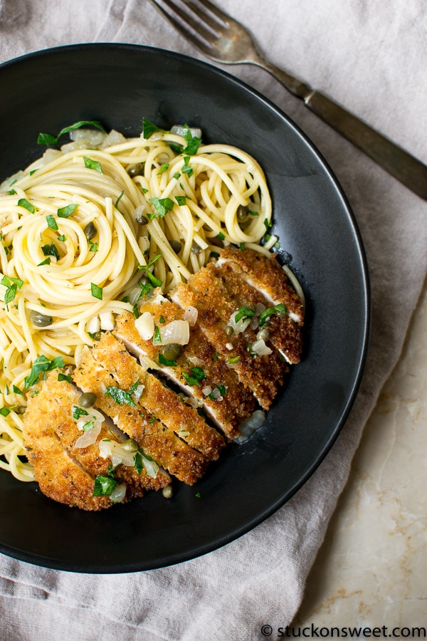 Golden crispy breaded chicken cutlet on a black plate with spaghetti and fresh herbs, served with capers and onions, delicious comfort food for dinner or lunch.