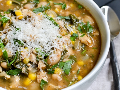 Creamy chicken and vegetable soup with beans, corn, kale, and shredded cheese, served in a white bowl with a black napkin and spoon, perfect for comfort food lovers.