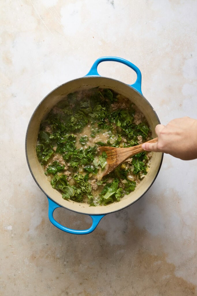 Creamy Zuppa Toscana soup with kale, sausage, and potatoes in a blue pot, being stirred with a wooden spoon. Perfect for comfort food lovers.