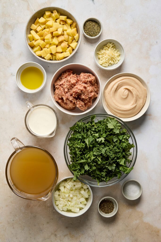 Ingredients for Zuppa Toscana soup including potatoes, kale, sausage, cream, and spices arranged on a light surface.