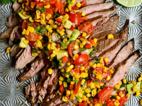 Juicy grilled steak sliced and topped with a fresh corn tomato salsa, garnished with lime wedges and cilantro on a decorative plate. Perfect for summer meals and steak dinner ideas.