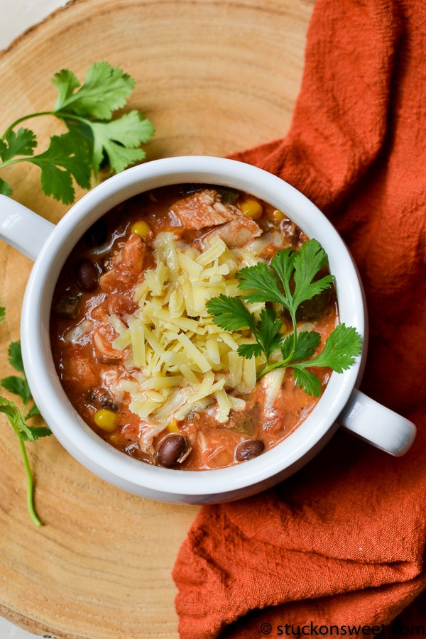 Creamy chicken chili with shredded cheese, black beans, corn, and fresh cilantro in a white mug, served on a wooden surface with a rustic orange cloth for a comforting meal.