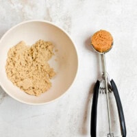 Golden brown sugar being measured with a stainless steel scoop on a white textured background for baking and dessert recipes.