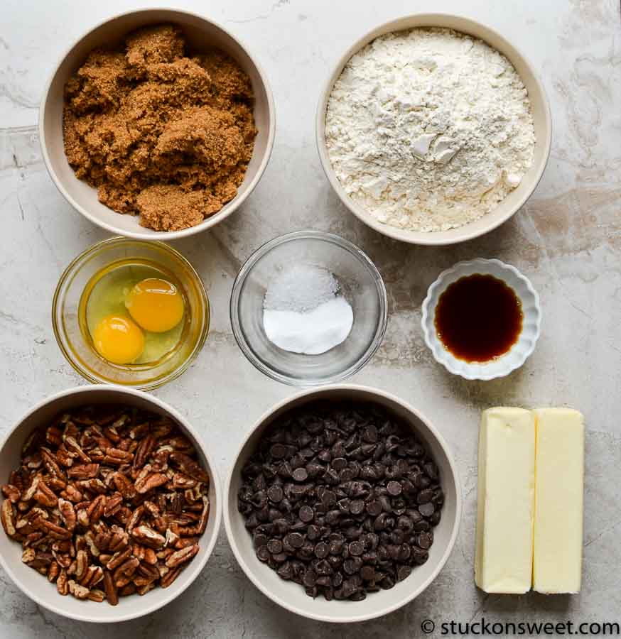 Brown sugar and chocolate chips for baking, with eggs, flour, vanilla, butter, salt, and pecans on a marble countertop. Perfect ingredients for homemade cookies or desserts.