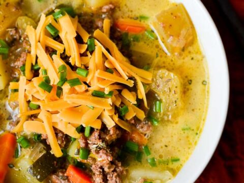 Creamy beef and vegetable soup topped with shredded cheddar cheese, fresh chives, and baked potatoes, served in a white bowl for a comforting and hearty meal.