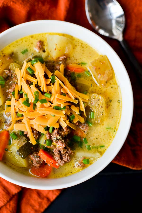 Creamy beef and vegetable soup topped with shredded cheddar cheese, fresh chives, and baked potatoes, served in a white bowl for a comforting and hearty meal.