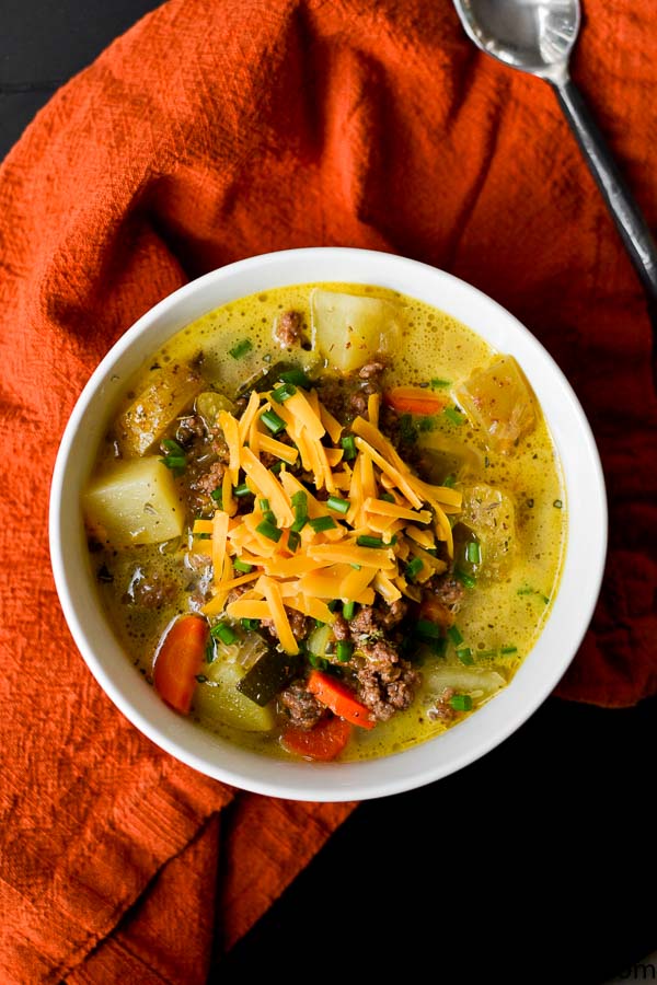 Creamy ground beef and vegetable soup topped with shredded cheddar cheese, served in a white bowl on an orange napkin. Hearty comfort food with fresh ingredients and savory flavors.