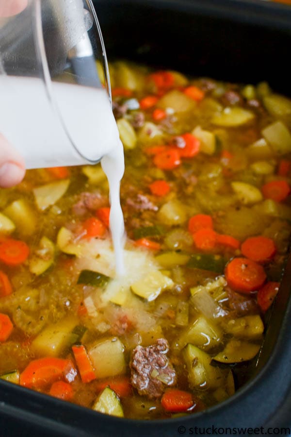 Creamy liquid being poured into a slow cooker filled with vegetables and ground beef, preparing a hearty stew.