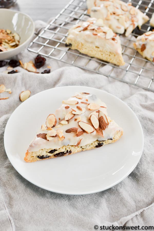Butter cake slice with almond topping, blueberry filling, white glaze, and sliced almonds on white plate.
