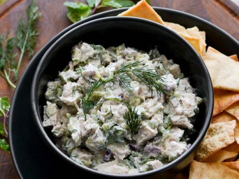 Creamy chicken salad with herbs and fresh dill in a black bowl, served with crispy pita chips on a wooden platter, perfect for snack or appetizer.
