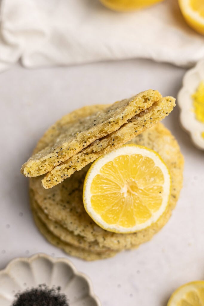 Lemon poppy seed cookies stacked with lemon slices and baking ingredients on a white surface.