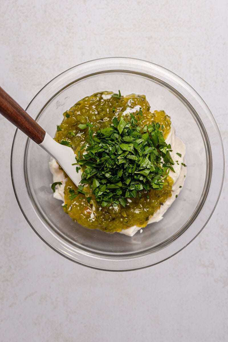 Chopped fresh herbs with green relish on a creamy white sauce in a glass mixing bowl for recipe preparation.