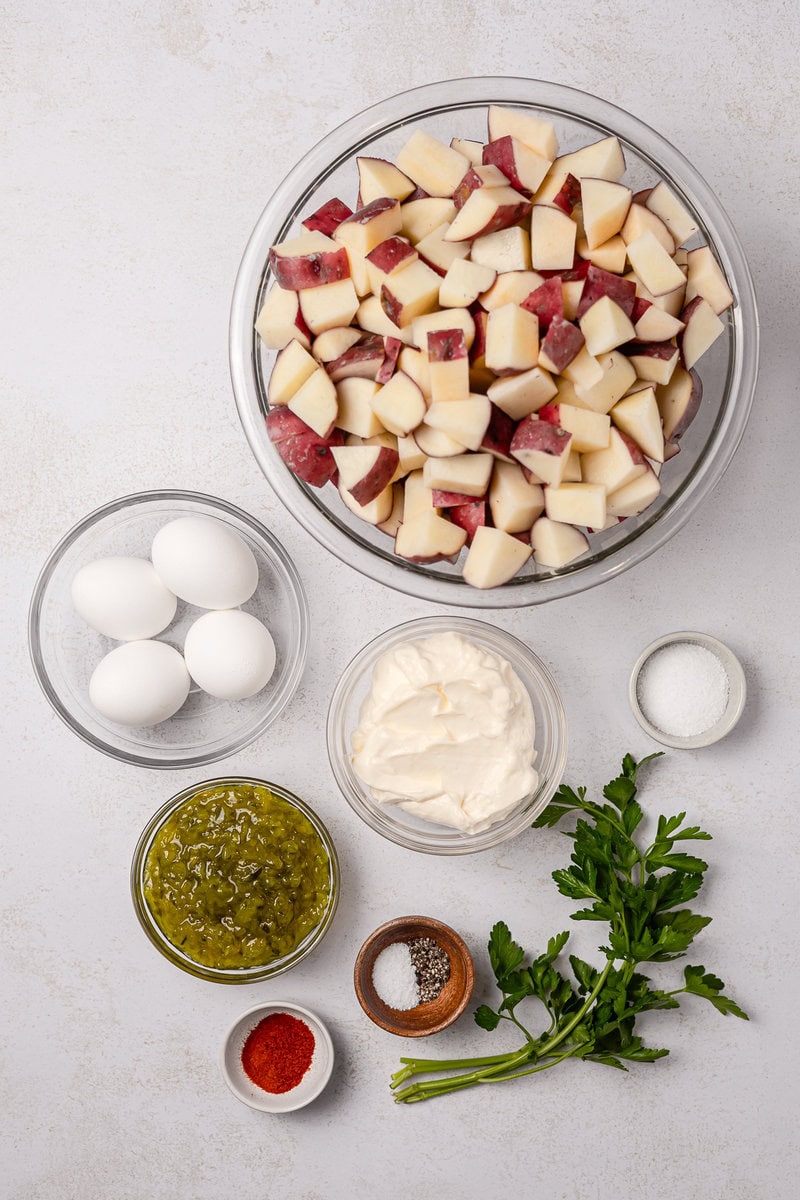 Diced red and white apples in a glass bowl, along with ingredients like eggs, cream cheese, chopped pickle relish, herbs, and spices for apple salad recipes.