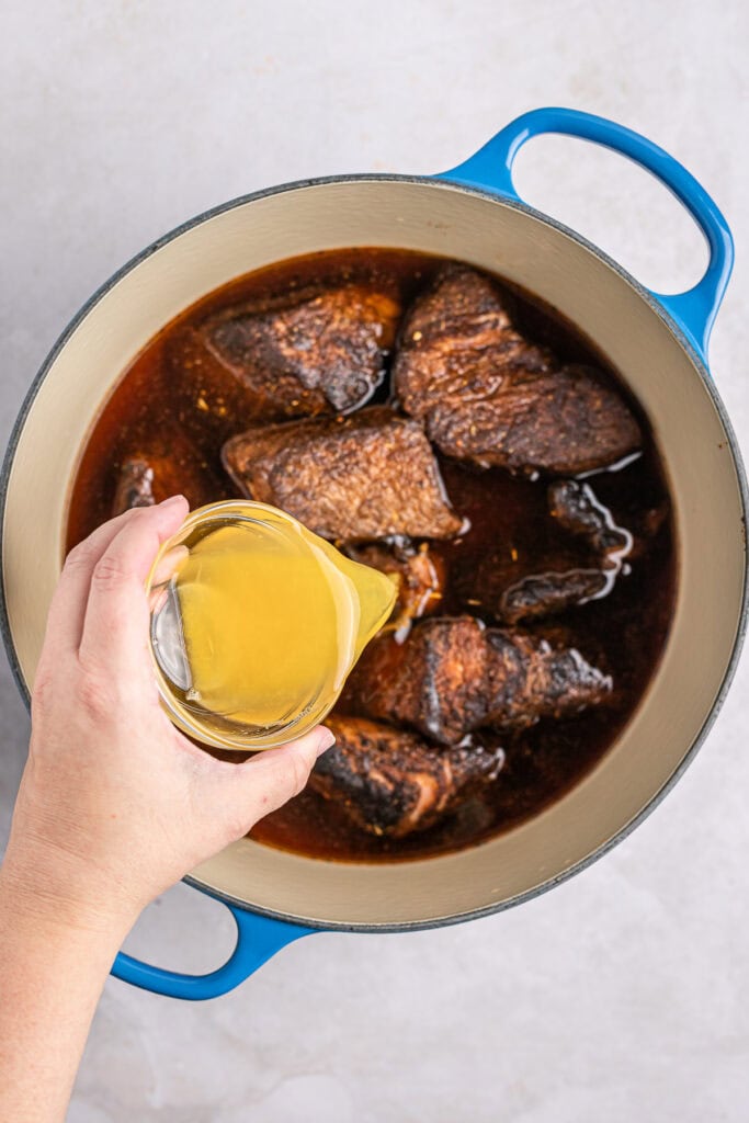Delicious homemade beef stew simmering in a white and blue ceramic pot, with a hand pouring a golden liquid, likely broth or oil, into the rich, savory meat stew, perfect for cold weather comfort food.