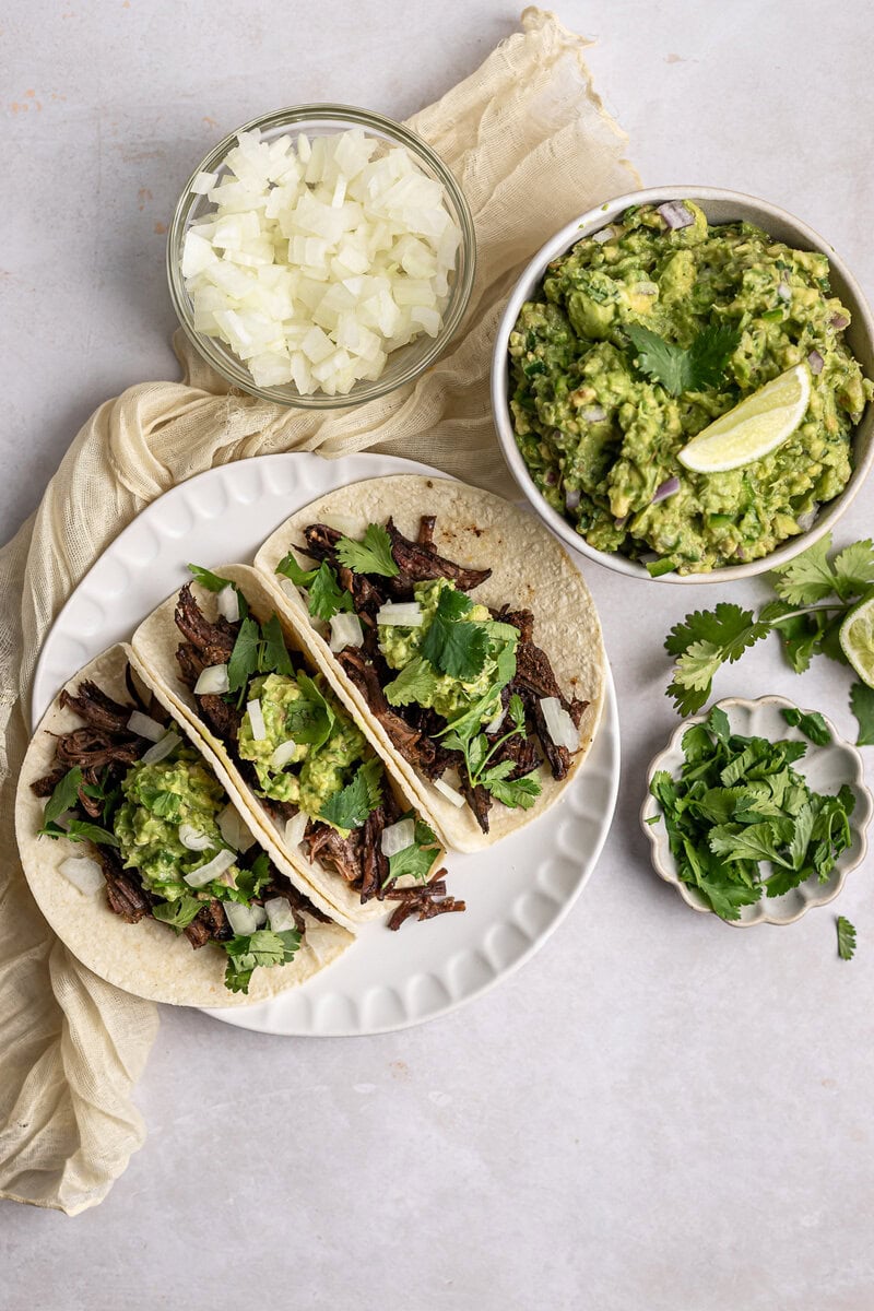 Creamy guacamole and shredded beef tacos on a white plate with chopped onions and cilantro, topped with fresh cilantro leaves, lime wedges, and onion in the background. Perfect for Mexican cuisine.