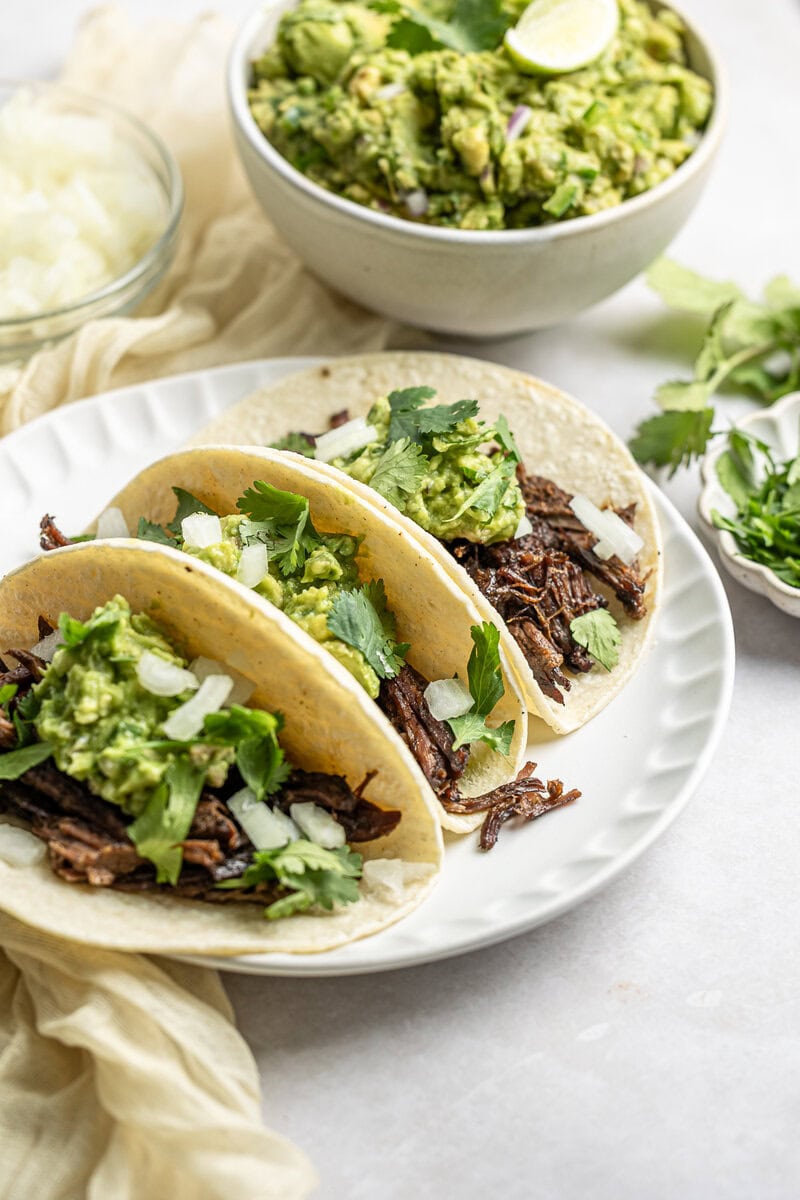 Creamy shredded beef tacos with fresh cilantro, guacamole, and onion on soft corn tortillas, served with a side of avocado and lime in a cozy, bright setting.