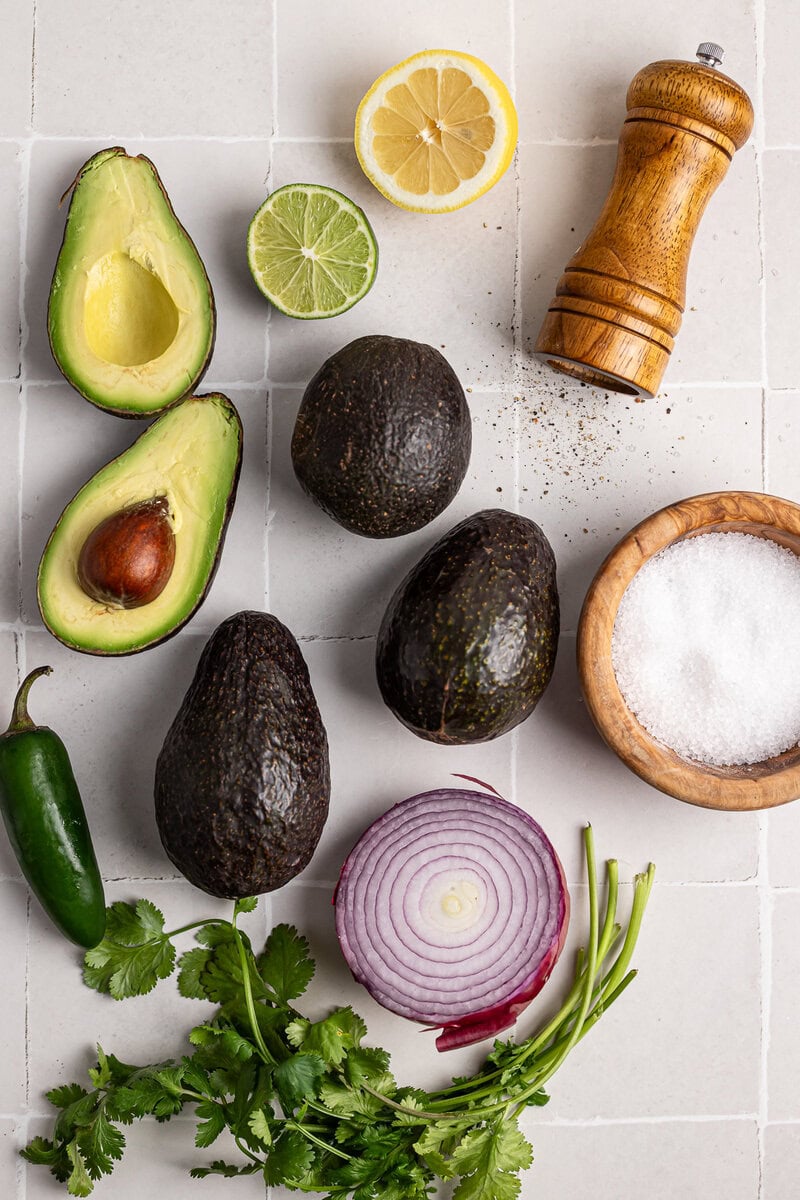Fresh avocados, lime, lemon, red onion, cilantro, sea salt, black pepper, and a pepper grinder on white tile background for healthy Mexican cuisine or guacamole preparation.