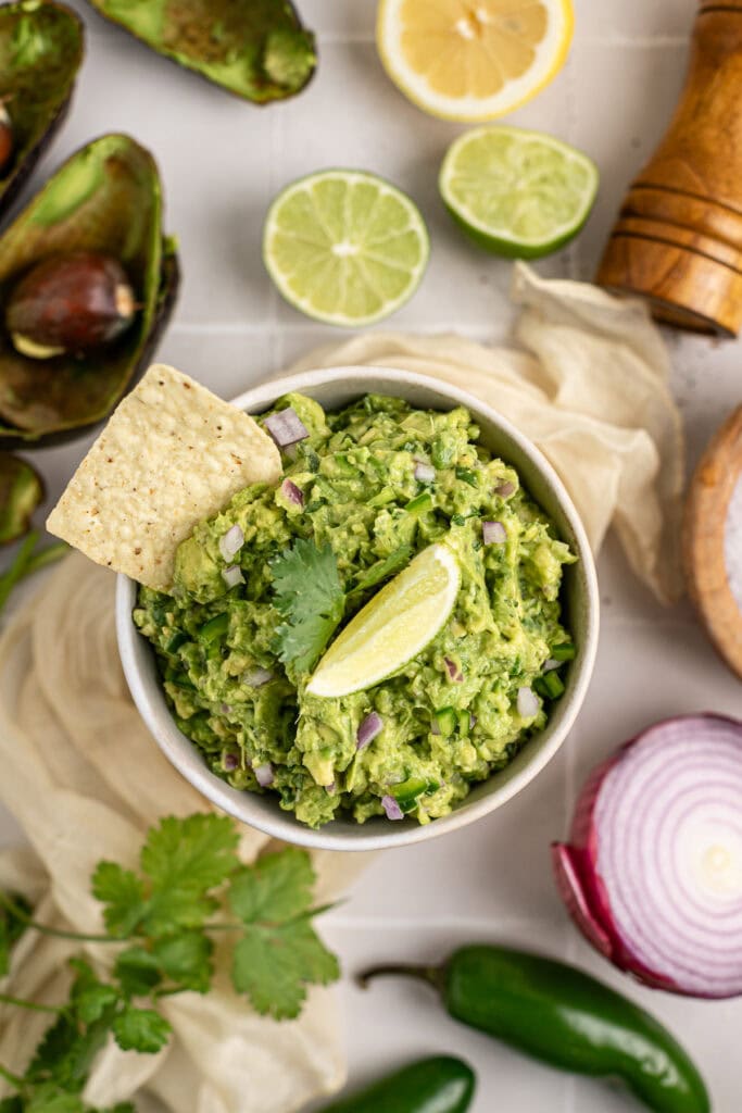 Creamy avocado guacamole topped with cilantro and red onions, served with lime wedges and tortilla chips, perfect for healthy snacking and party appetizers.