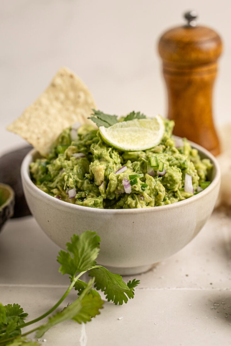 Creamy avocado salad garnished with lime wedge and cilantro in a white bowl. Topped with red onion and served with crispy tortilla chips, perfect for healthy snacking and guacamole recipes.