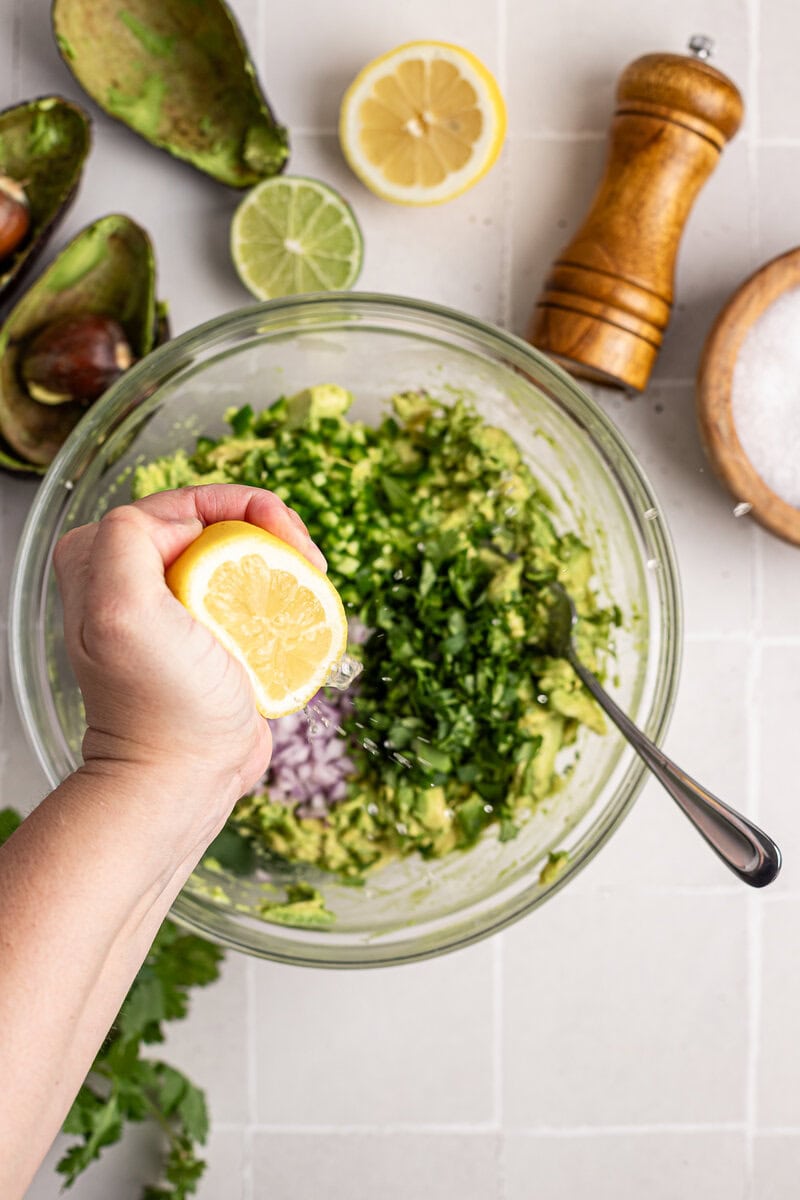 Juice being squeezed from a lemon into a bowl of chopped avocado, onion, and cilantro for guacamole preparation.