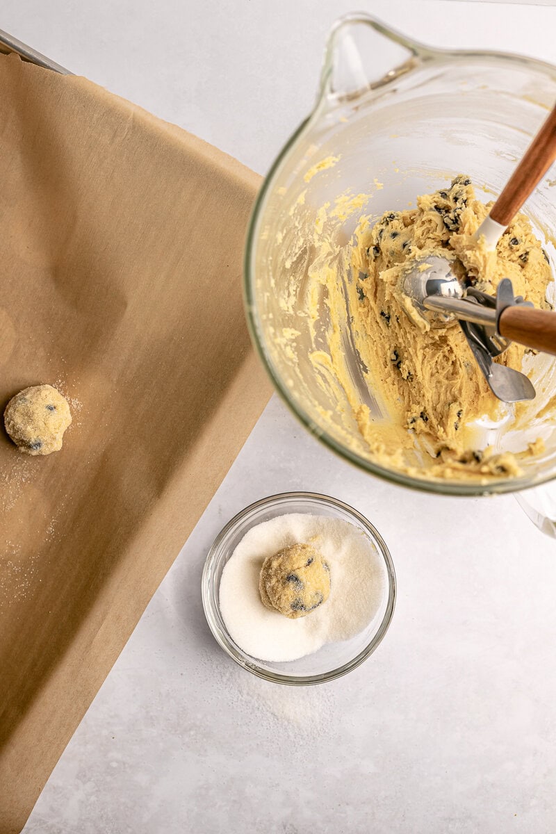 Creamy cookie dough mixing in a glass bowl with chocolate chips, ready to be baked into delicious cookies. Cookie dough balls coated in sugar on parchment paper for baking at Stuck On Sweet.