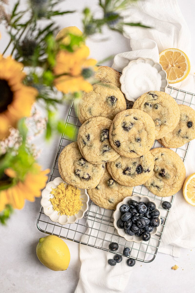 Delicious homemade blueberry cookies on a cooling rack surrounded by lemons, blueberries, and lemon zest, perfect for sweet tooths and cookie lovers.
