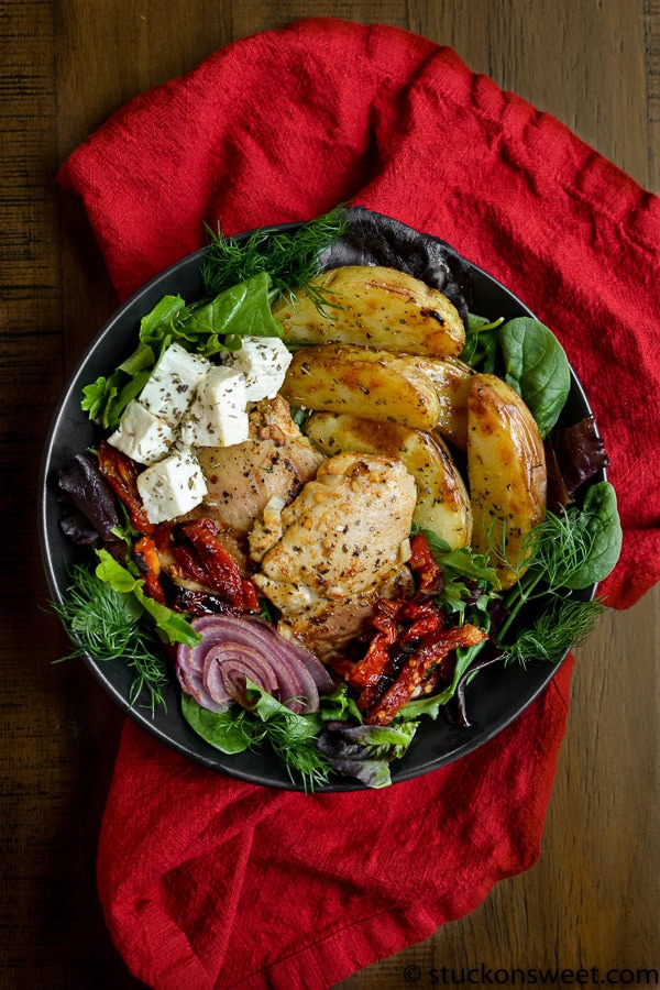 Fresh Greek salad with grilled chicken, roasted potatoes, feta cheese, sun-dried tomatoes, and mixed greens, served on a black plate with a red cloth background.
