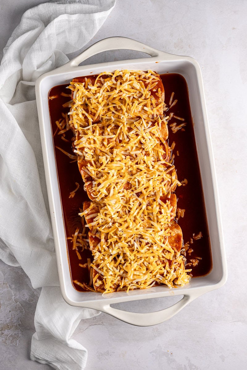 Baked cheesy enchiladas with shredded cheese topped with enchilada sauce in a white baking dish.