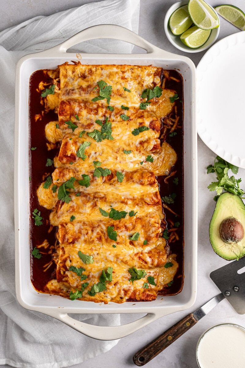 Creamy Chicken Enchil casserole with melted cheese, savory sauce, fresh cilantro, and avocado slices, served with lime wedges, perfect for a delicious Mexican-inspired meal.