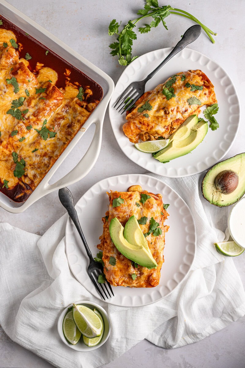 Cheesy chicken enchiladas with avocado slices on white plates, garnished with cilantro and lime wedges, served alongside baked enchiladas with melted cheese in a casserole dish.