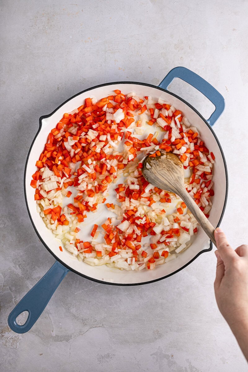 Diced onions and red bell peppers cooking in a white skillet, being stirred with a wooden spoon, on a gray countertop.