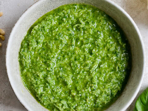 Creamy pesto sauce with fresh herbs in a white bowl, lemon wedge, grated cheese, and pine nuts on light background for culinary and recipe content.