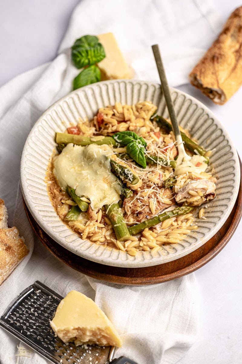 Creamy baked pasta with melted cheese, fresh basil, and tender asparagus in a rustic bowl, served with garlic bread and Parmesan cheese on a cozy white cloth. Perfect comfort food for family dinners.