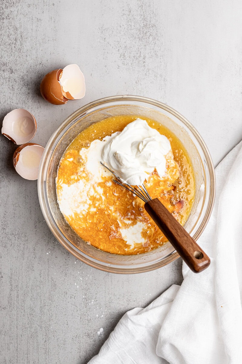 Creamy mixing bowl with eggs, sour cream, and vanilla for cake batter, with cracked eggshells nearby, on a gray surface.