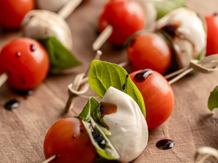 Bright caprese skewers with cherry tomatoes, mozzarella, fresh basil, and balsamic glaze on a wooden serving board. Perfect for Mediterranean-inspired appetizers and party snacks.