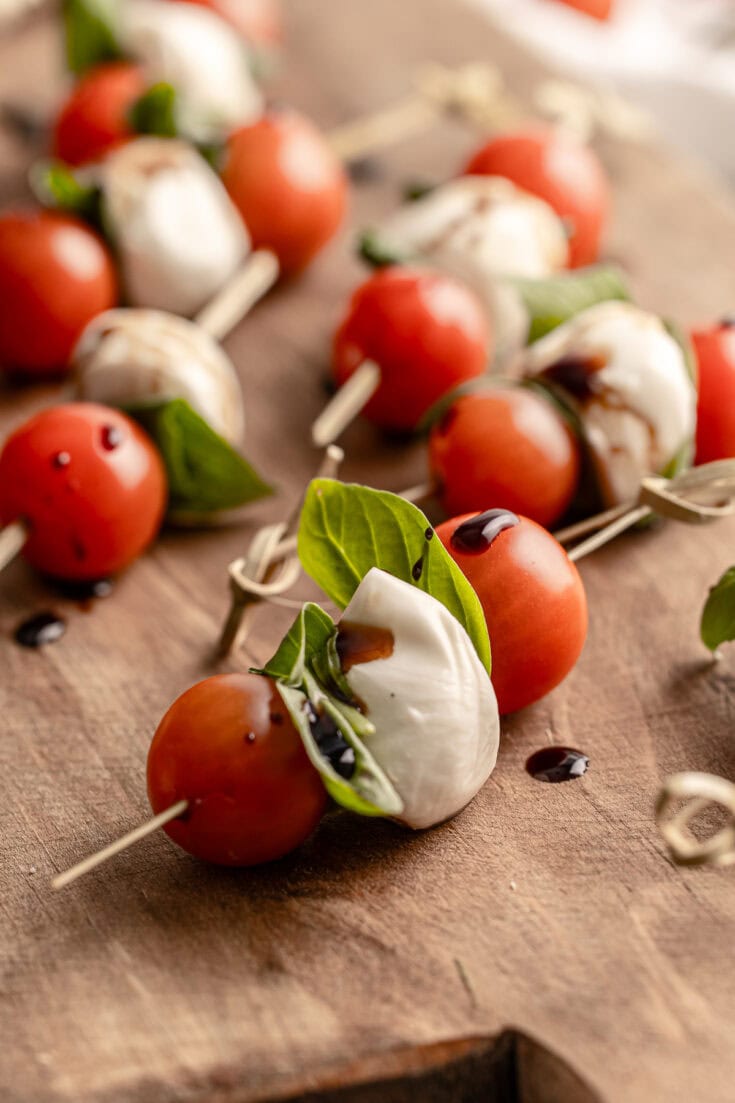 Bright caprese skewers with cherry tomatoes, mozzarella, fresh basil, and balsamic glaze on a wooden serving board. Perfect for Mediterranean-inspired appetizers and party snacks.
