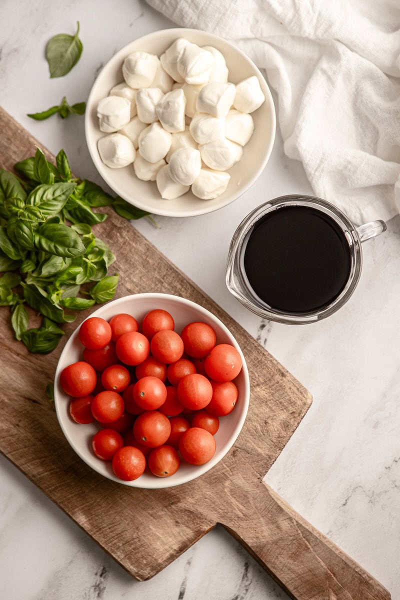 Fresh cherry tomatoes, fresh basil, mozzarella balls, and balsamic vinegar on a wooden cutting board, ideal for Caprese salad ingredients.