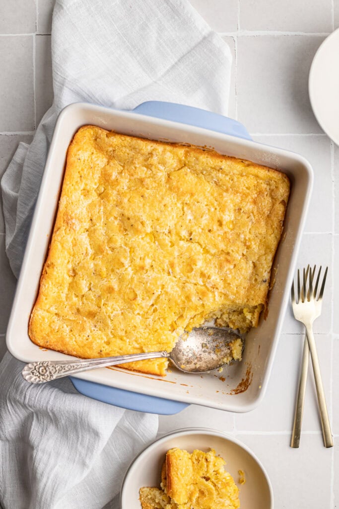 Corn Casserole with golden edges in a white baking dish. Freshly served with a piece on a small plate and a vintage spoon, perfect for sweet tooth cravings or dessert recipes.