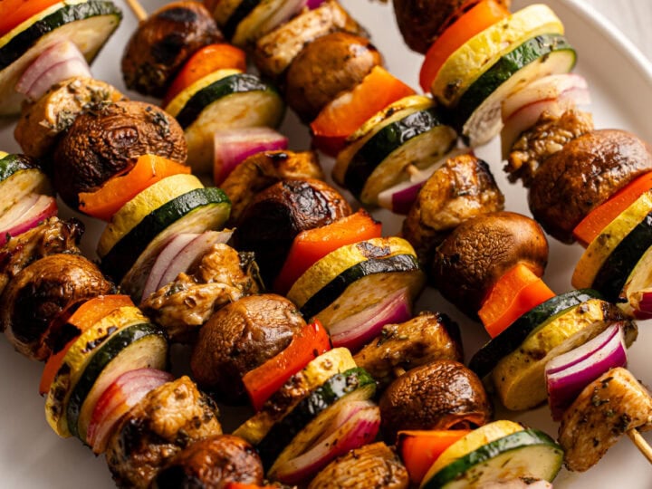 Colorful grilled vegetable skewers with mushrooms, zucchini, bell peppers, and red onions on a white plate for healthy, delicious outdoor grilling recipes, Vegetarian skewers, Summer BBQ ideas.