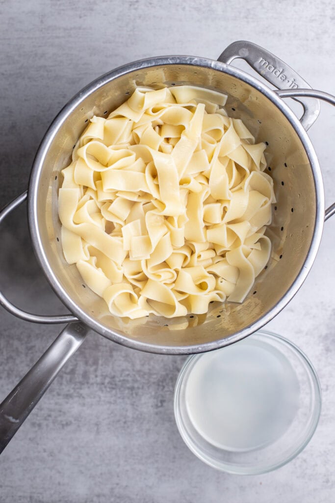 Creamy homemade pasta noodles in a stainless steel colander with a glass of milk on the side, perfect for delicious Italian-inspired recipes.