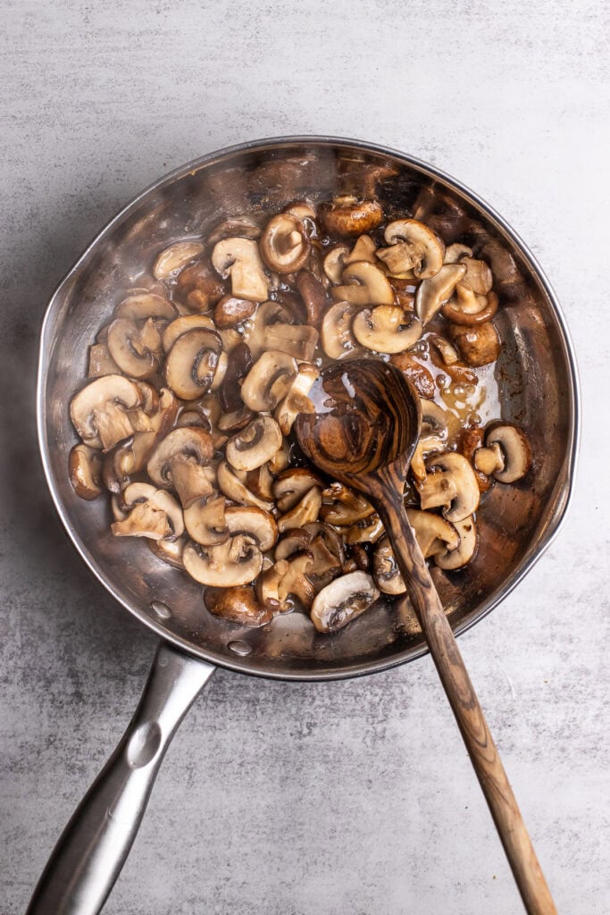 Sautéed mushrooms in a stainless steel skillet with a wooden spoon, fresh and cooked mushroom slices, cooking process, perfect for mushroom recipes.