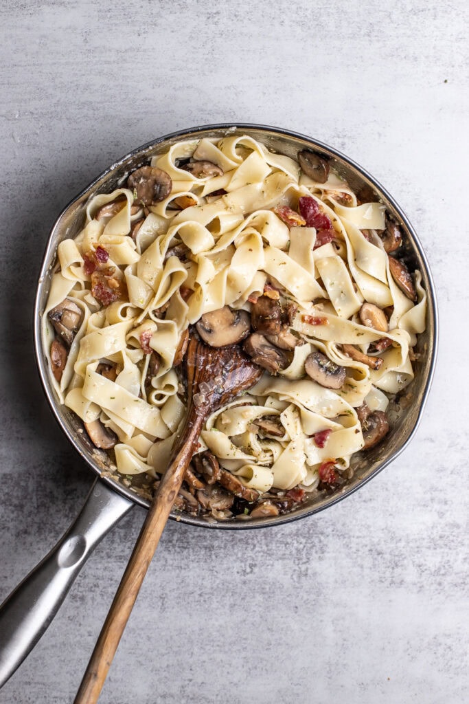 Creamy mushroom and bacon fettuccine pasta in a stainless steel skillet with a wooden spoon.
