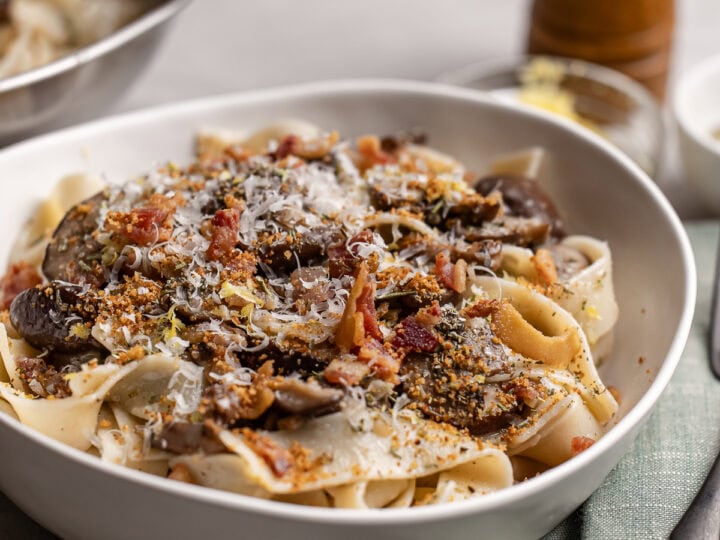 Creamy mushroom bacon pasta with grated cheese and fresh herbs, served in a white bowl on a rustic wooden table; perfect comfort food with savory flavors and crispy bacon accents.