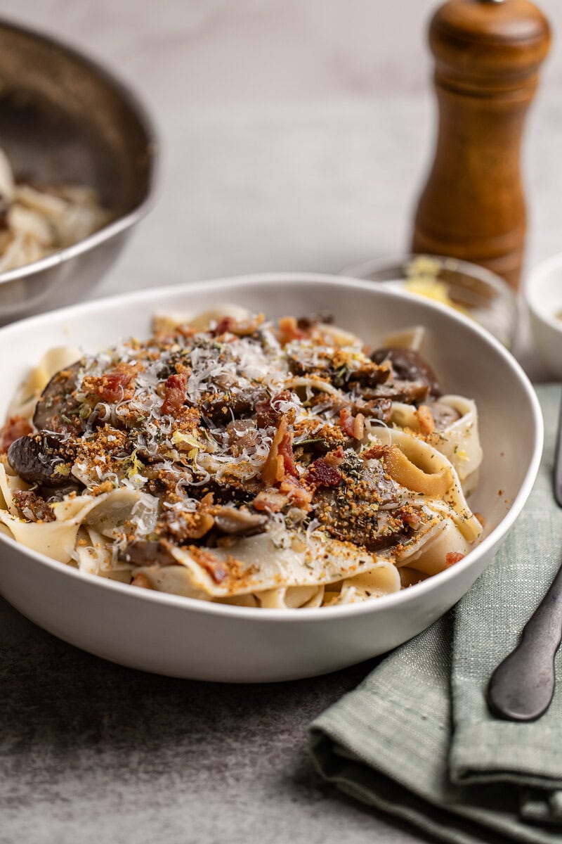 Creamy mushroom bacon pasta with grated cheese and fresh herbs, served in a white bowl on a rustic wooden table; perfect comfort food with savory flavors and crispy bacon accents.