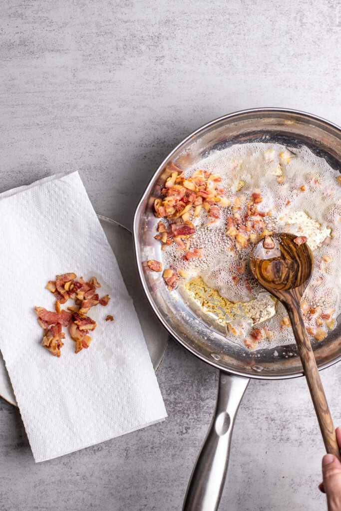 Crispy bacon being cooked in a stainless steel skillet on a gray surface with paper towels nearby.
